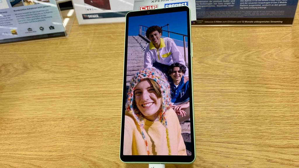Ein getestetes Smartphone mit 6 Zoll liegt eingeschaltet mit einem Foto von Personen auf einer Holzfläche.