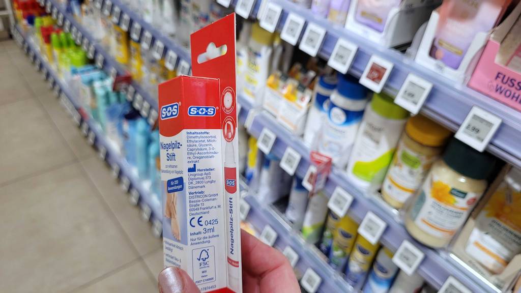 Nagelpilzlack-Test: Eine Hand zeigt die Seite einer Verpackung, auf der Informationen zum Inhalt und Hersteller stehen.