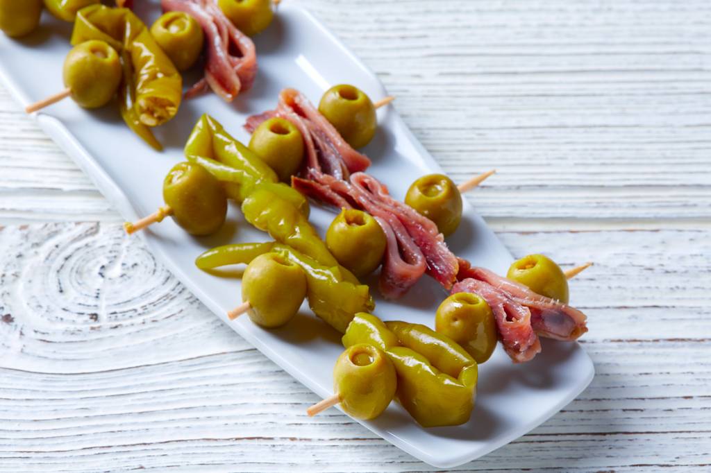 Spanische-Oliven-Test: Auf einem langen Teller befinden sich Oliven, grüne Paprika und Anchovis. Diese sind auf einem Zahnstocher aufgespießt. 