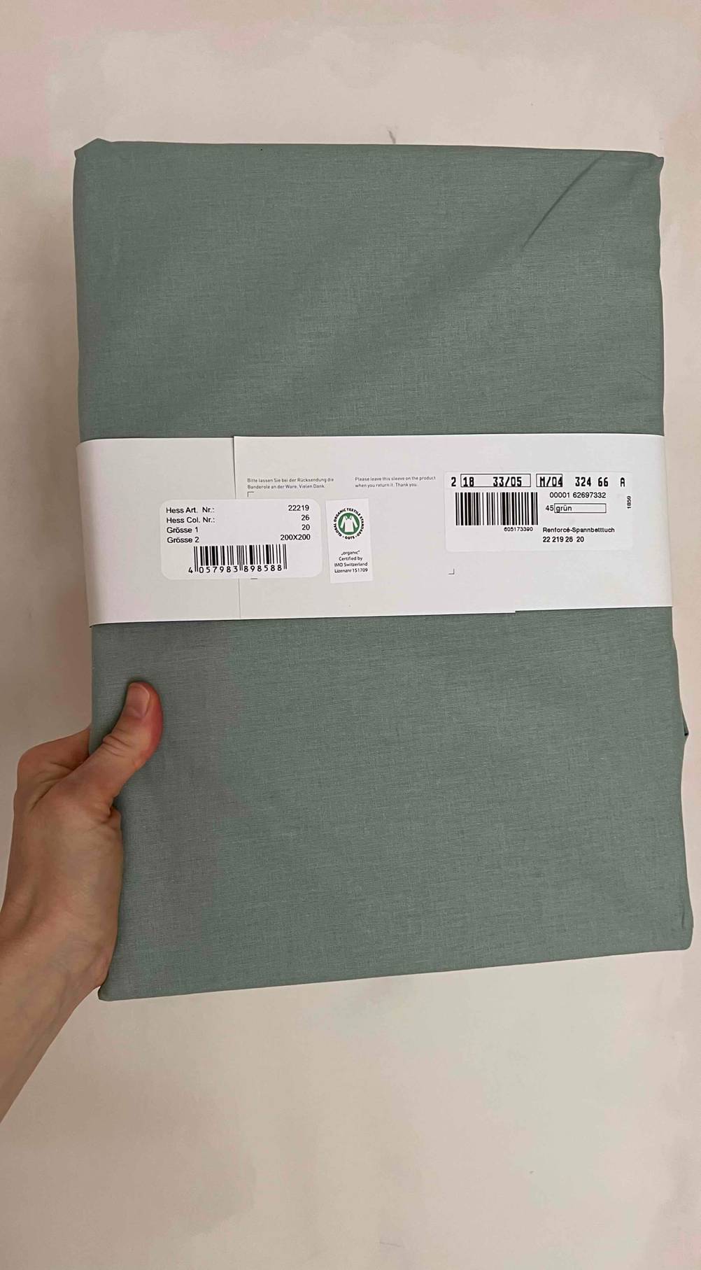 Spannbettlaken-180x200-Test: Eine Hand hält ein verpacktes Spannbettlaken mit der Rückseite vor eine helle Wand.