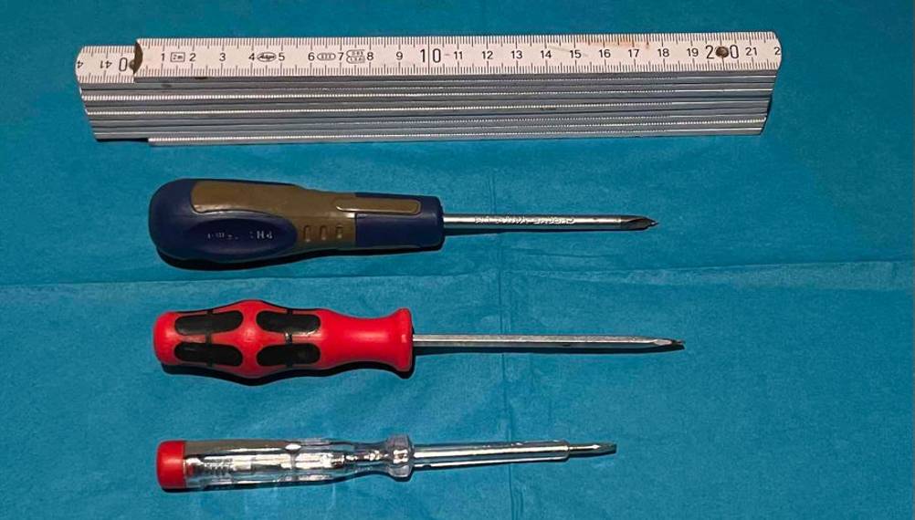 Spannungsprüfer-Schraubendreher im Test liegt neben zwei weiteren Schraubendrehern und einem weißen Zollstock auf einem türkisfarbigen Untergrund.