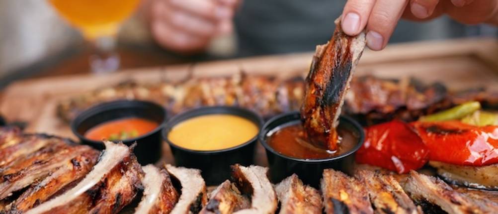 Spareribs-Halter im Test: Eine Hand taucht Spareribs in verschiedene Saucen. Mehrere gegarte Spareribs liegen im Vordergrund.