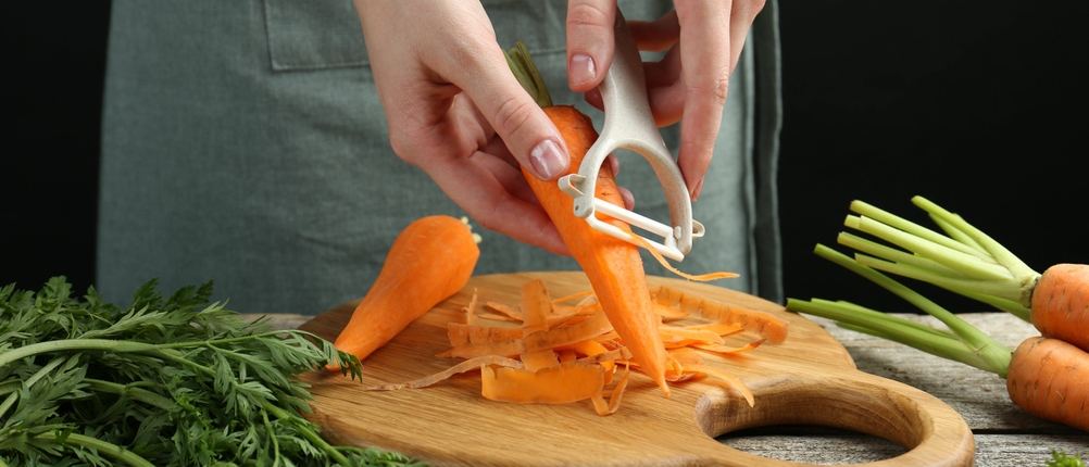 Karotten werden mit einem Sparschäler auf einem Holzbrett geschält.