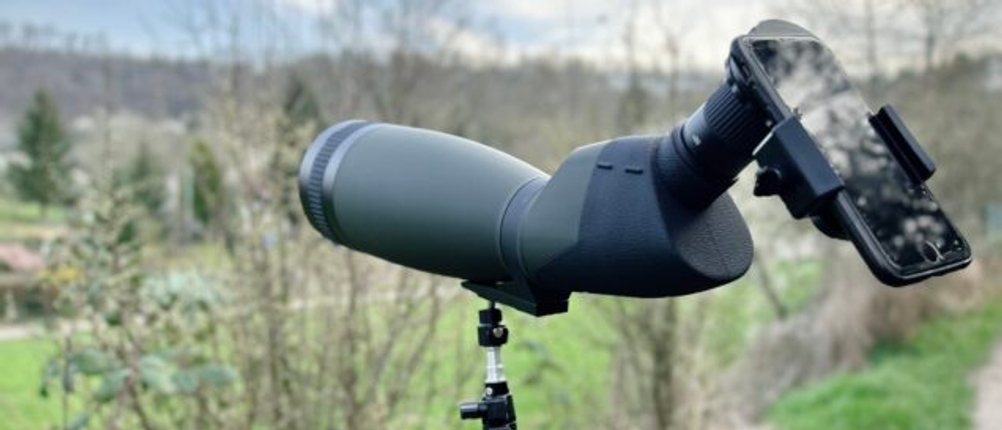 Das Bresser Travel 20-60x60 Spektiv steht auf einem Stativ und ist mit einem Smartphone-Adapter für Digiscoping verbunden. Im Hintergrund ist eine Wiese mit Bäumen sichtbar.