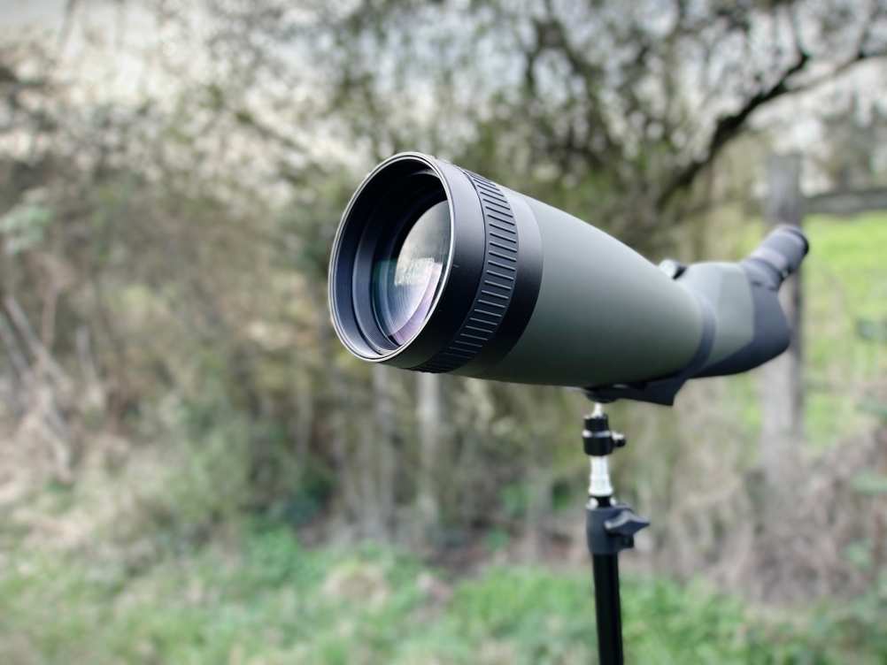 Frontale Nahaufnahme der Objektivseite des Bresser Travel 20-60x60 Spektivs auf einem Stativ. Die große Linse spiegelt Licht und Bäume wider.