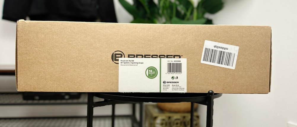 Der Versandkarton des Bresser Travel 20-60x60 Spektivs steht auf einem schwarzen Tisch. Das Etikett mit Barcode und Produktinformationen ist gut sichtbar.