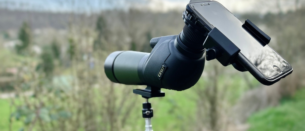 Das Gosky 20-60x60 Spektiv ist auf einem Stativ befestigt und mit einem Smartphone für Digiscoping verbunden. Im Hintergrund ist eine Wiesenlandschaft zu sehen.