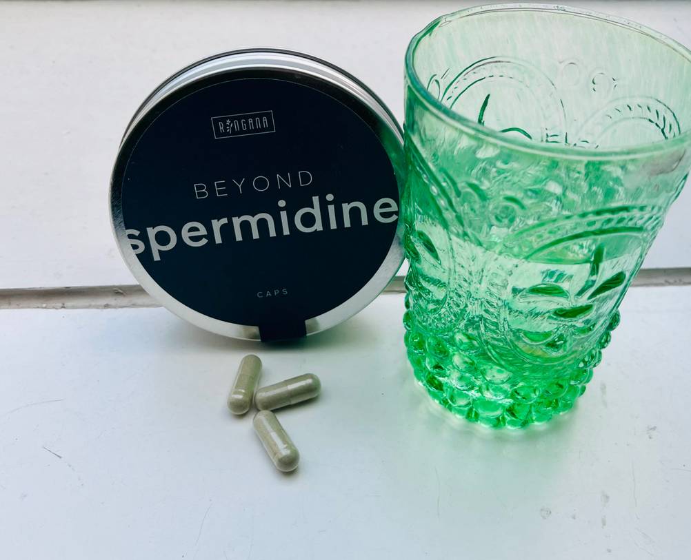 Neben einem Glas Wasser steht eine Dose Spermidin-Kapseln im Test und davor drei Kapseln auf einer hellen Fläche.