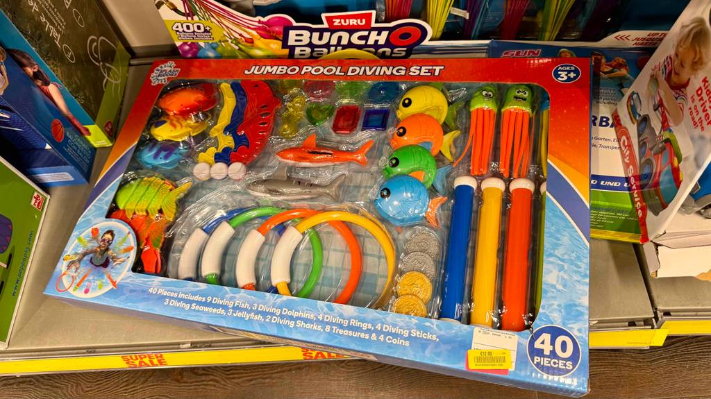 Das Tauchspielzeug im Test setzt sich aus 40 bunten Einzelteilen zusammen, die innerhalb der Verpackung auf einem Verkaufsregal neben anderem Spielzeug liegt.