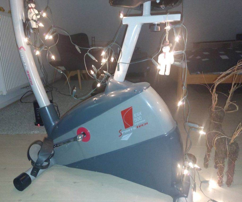 Ergometer-Test: Seitliche Nahaufnahme eines blauen Modells ESX 500 der Marke Sportstech, mit einer leuchtenden Lichterkette geschmückt, in einem Schlafzimmerbereich.