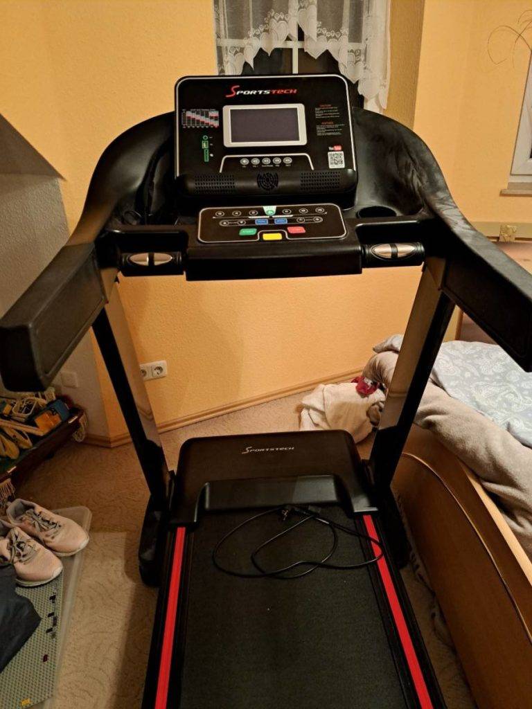 Ein schwarzes, getestetes klappbares Laufband der Marke Sportstech steht in einem Schlafzimmerbereich, mit Fokus auf die Halterung inklusive ausgeschaltetem LCD-Bildschirm sowie verschiedene Einstelltasten.