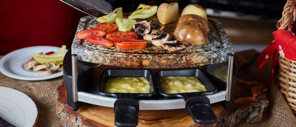 Spring-Raclette-Test: Auf einem Tisch steht ein Raclette mit Steinplatte und mit Käse befüllten Pfännchen. Die Steinplatte ist mit Tomaten, Kartoffeln und Champignons belegt. 