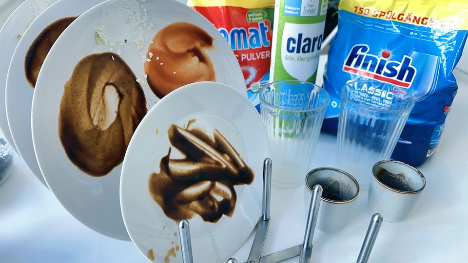 Mehrere verschmutzte Geschirrteile und Spülmaschinenpulver stehen auf einem Tisch.
