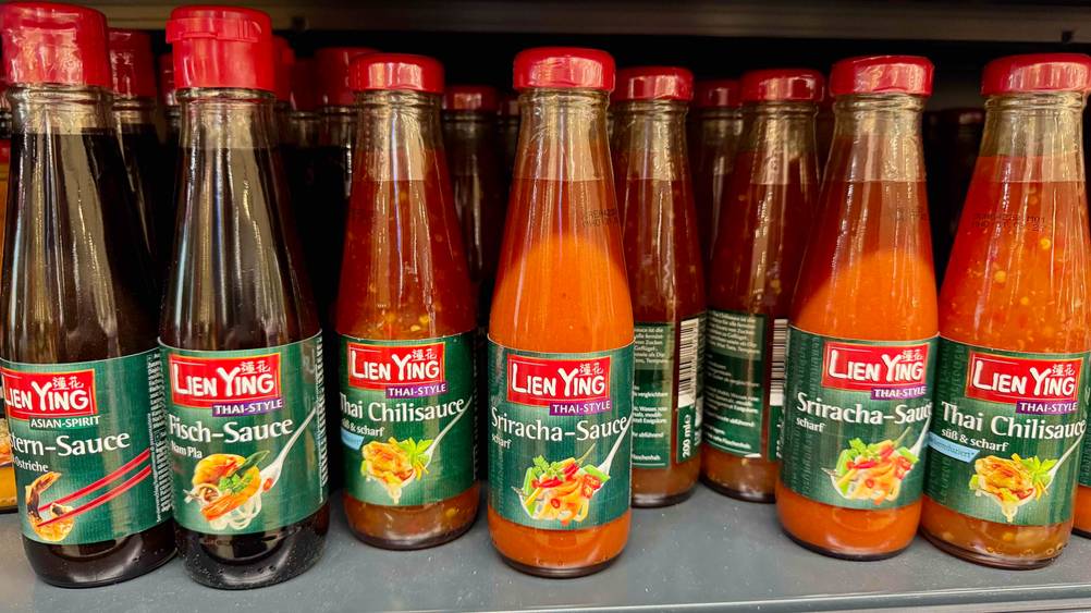 Mehrere Flaschen der getesteten Sriracha-Sauce in einem Verkaufsregal neben anderen Produkten.