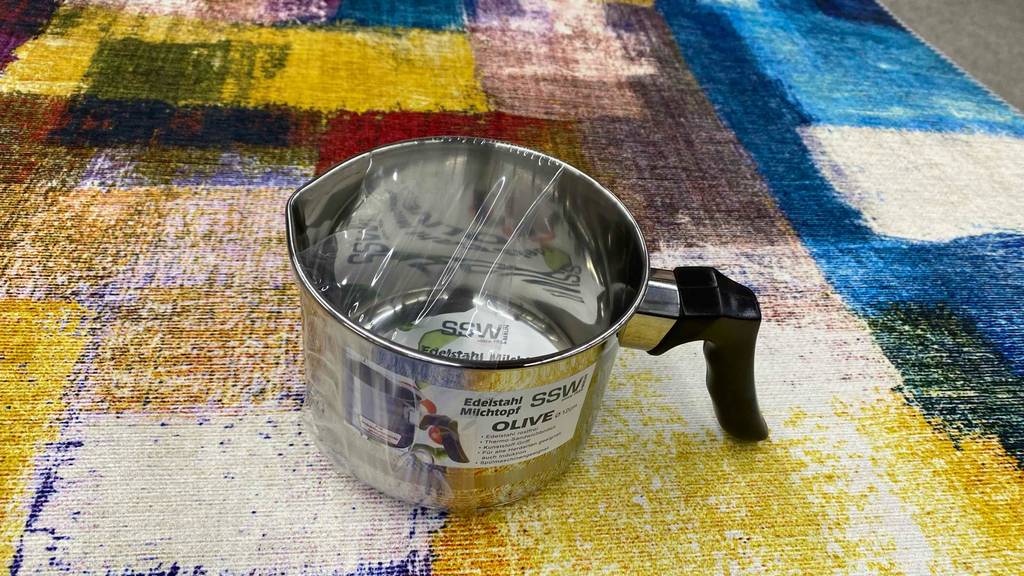 Im Test: ein Milchtopf aus Edelstahl mit schwarzem Kunststoffgriff steht in Cellophan eingepackt auf einer bunten Tischdecke.