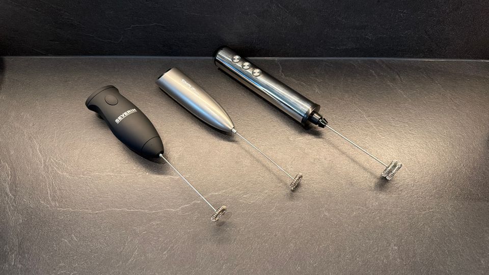 Drei Stab-Milchaufschäumer liegen nebeneinander auf einer Küchen-Arbeitsplatte.