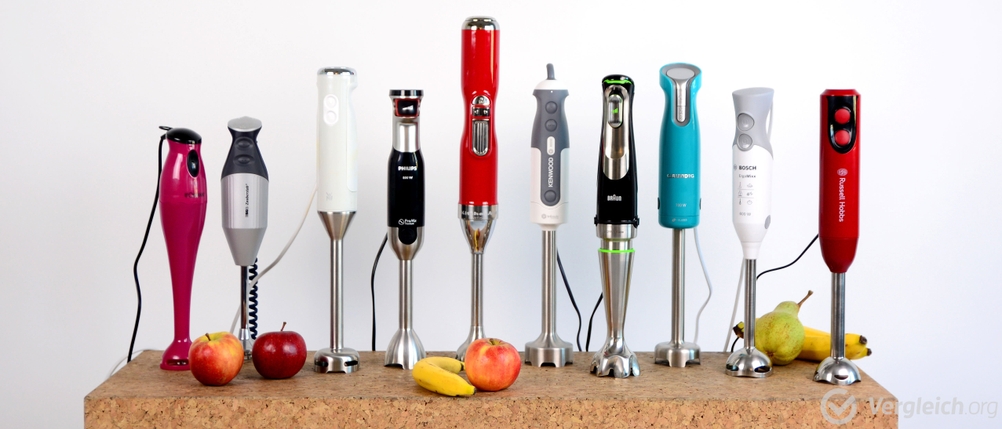 Mehrere Stabmixer stehen in einer Reihe, mit Obst im Vorder- und Hintergrund.