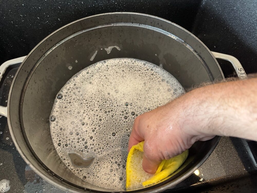 Der Behälter des Staub Cocotte 1007460 wird von einer Hand mit einem Lappen und Wasser gereinigt