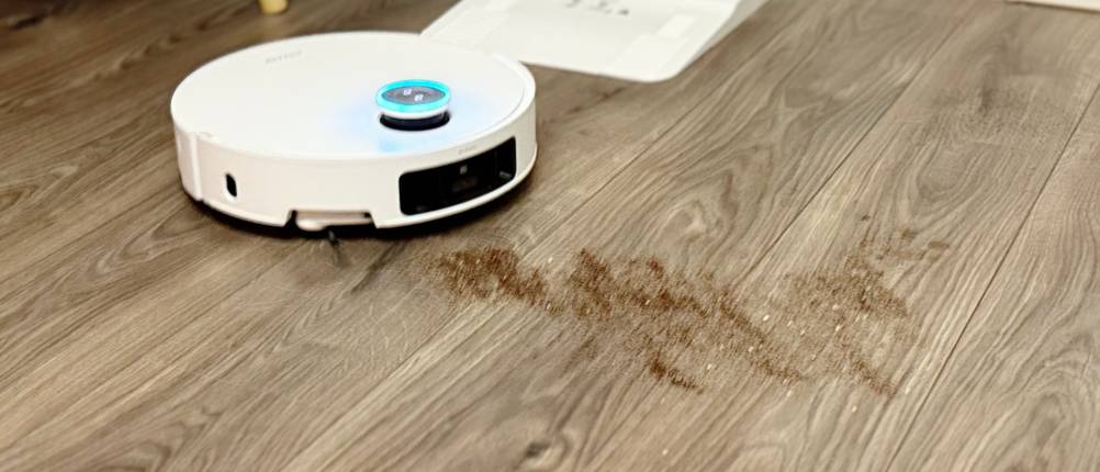 Ein weißer Staubsaugerroboter saugt Staub auf einem grauen Laminatboden auf.