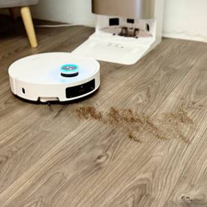 Ein weißer Staubsaugerroboter saugt Staub auf einem grauen Laminatboden auf.