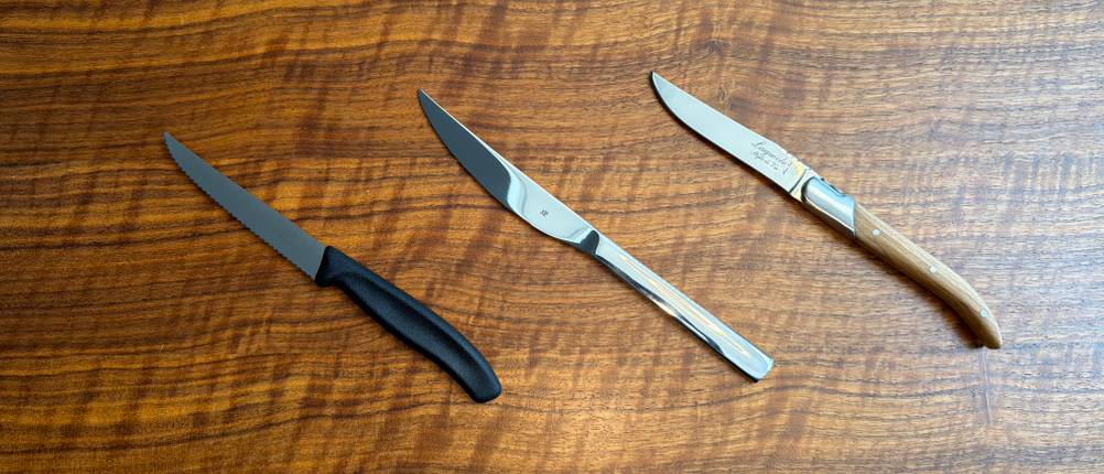 Drei Steakmesser liegen nebeneinander auf einem Holztisch