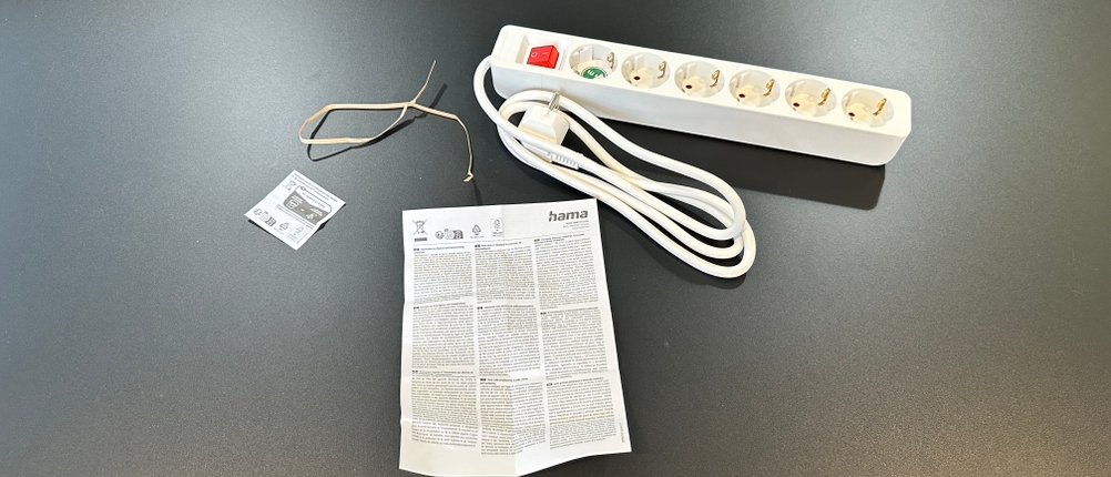 Die Hama-Steckdosenleiste und der zugehörige Lieferumfang liegen vollständig ausgelegt auf dem Tisch.