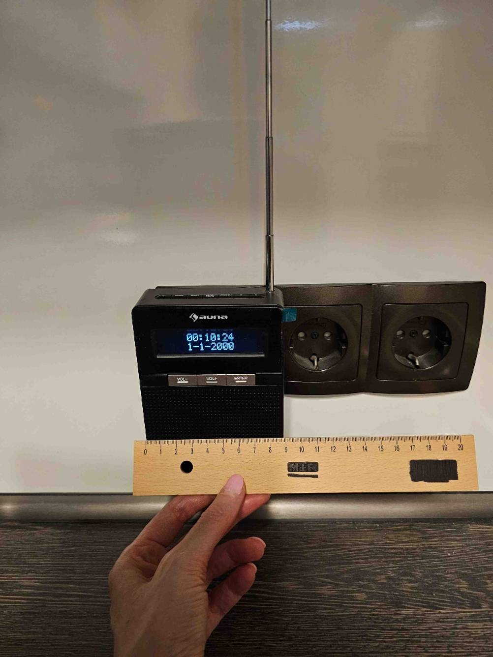 Steckdosenradio getestet: Ein Radio steckt in einer dunklen Steckdose, eine Hand hält ein Lineal an die untere Kante.