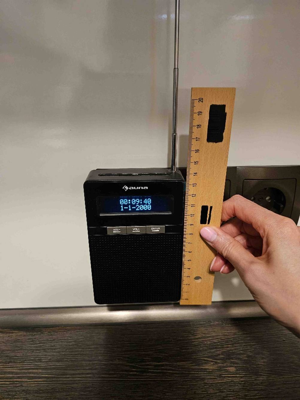 Ein getestetes Steckdosenradio steckt in einer Steckdosenleisete eine Hand hält ein Lineal an die rechte Seite.