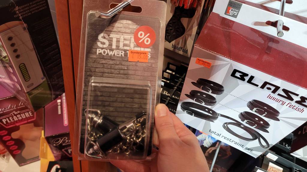 Nippelklemmen im Test: Hand hält Nippelklemmen der Marke Steel Power Tools zum Sonderpreis in ihrer Kunststoffverpackung eingeschweißt, die an einem Verkaufsstand mit anderen Sex-Toys hängt.