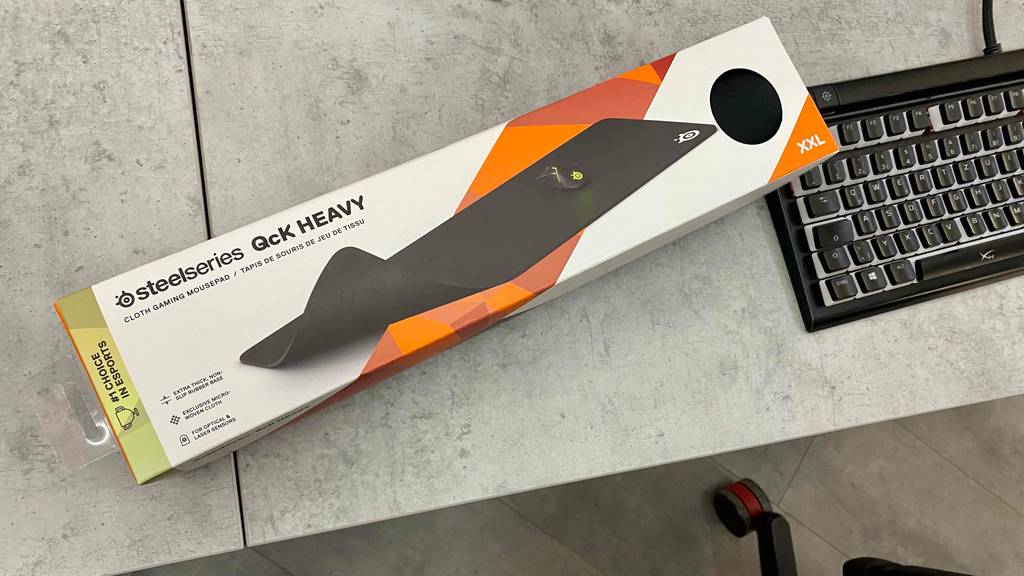 Eine weiß-orangefarbene Verpackung von einem Steelseries XXL-Mauspad im Test lehnt an einer schwarzen Tastatur auf einer hellgrauen Ausstellungs-Tischplatte.