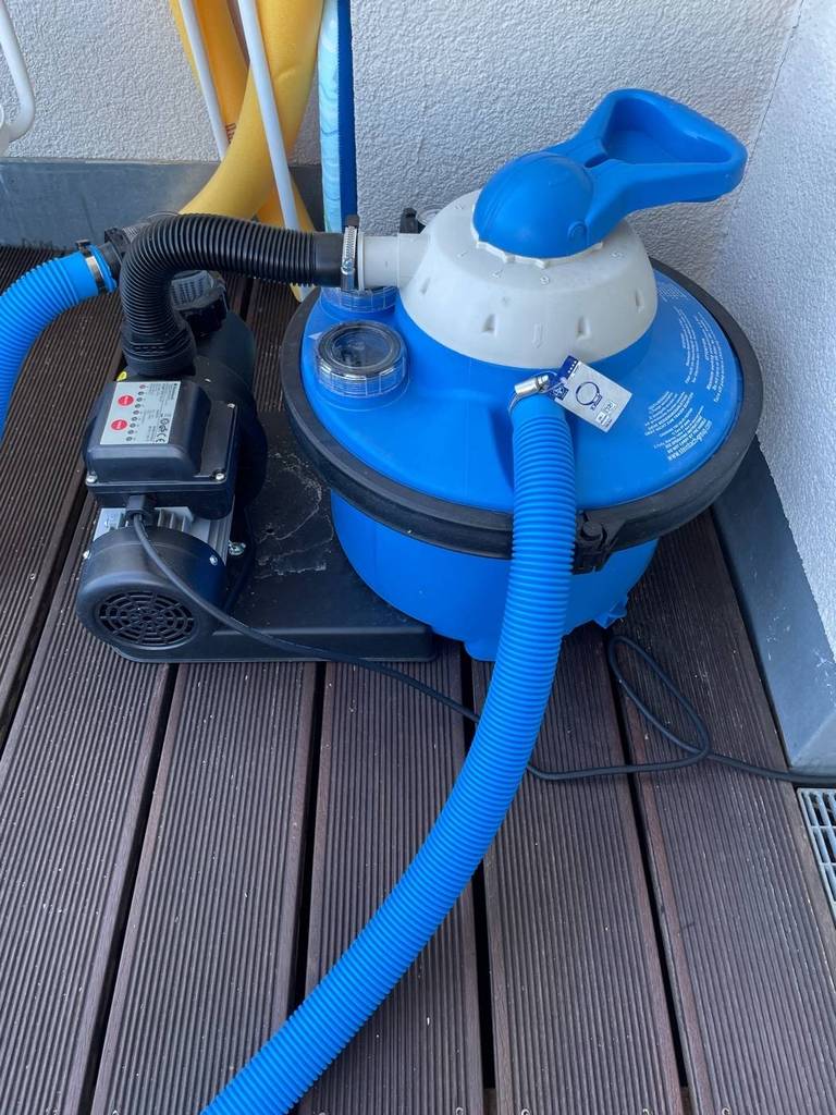 Getestete Sandfilteranlage in blau-schwarzem Design steht inklusive Schlauchsystem und Motor auf einem braunen Terrassenboden.