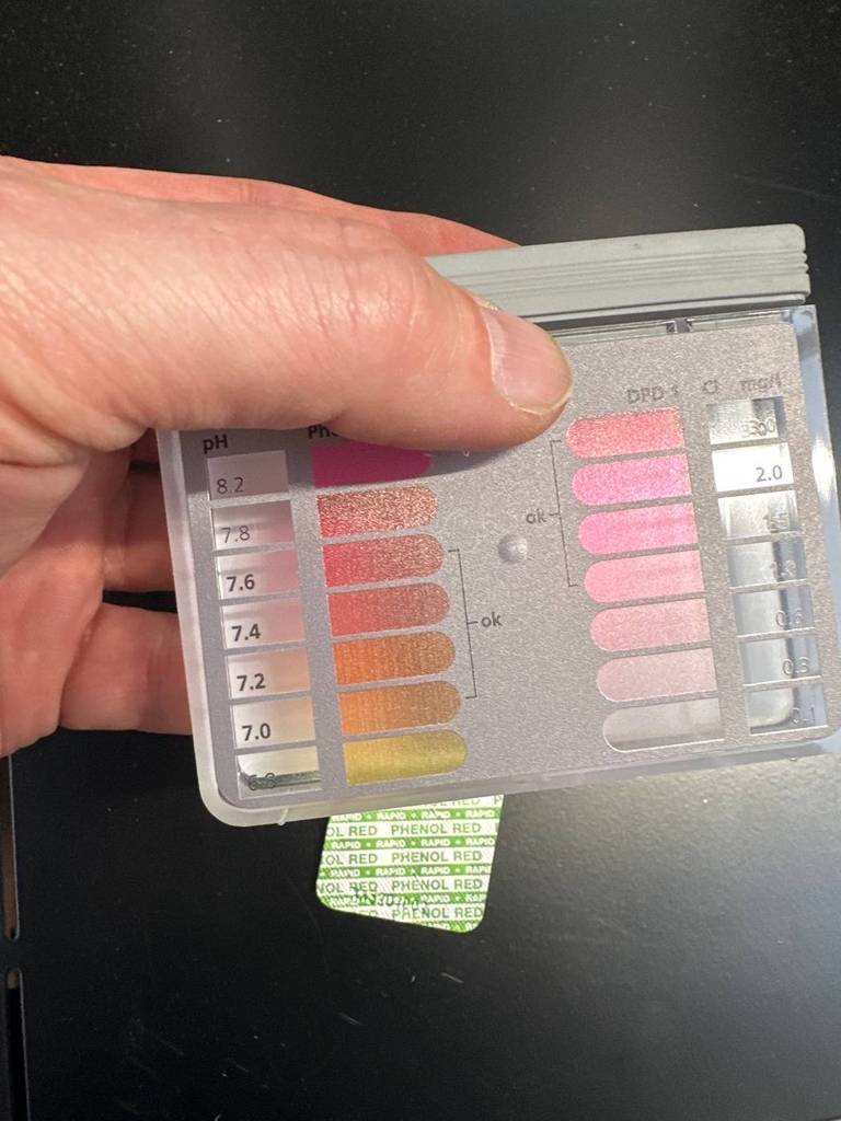 Pooltester-Test: Nahaufnahme einer Hand, die eine Hartplastik-Messtafel mit verschiedenen Farbbereichen und pH-Werten vor einen schwarzen Hintergrund hält.