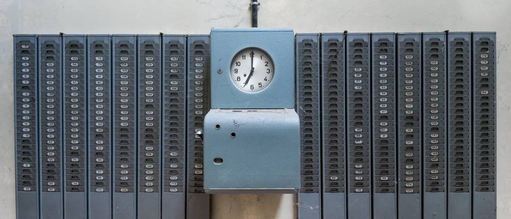 Stempeluhr-Test: Eine alte blaue analoge Stempeluhr an einer Wand mit Regalen für Stempelkarten.