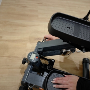 Eine Person greift mit einer Hand nach einer Einstellschraube in der Mitte zwischen den beiden Pedalen des Steppers.