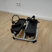 Ein kompakter grau-schwarzer Mini-Stepper mit rutschfesten Pedalen und Widerstandsseilen auf einem Holzboden.
