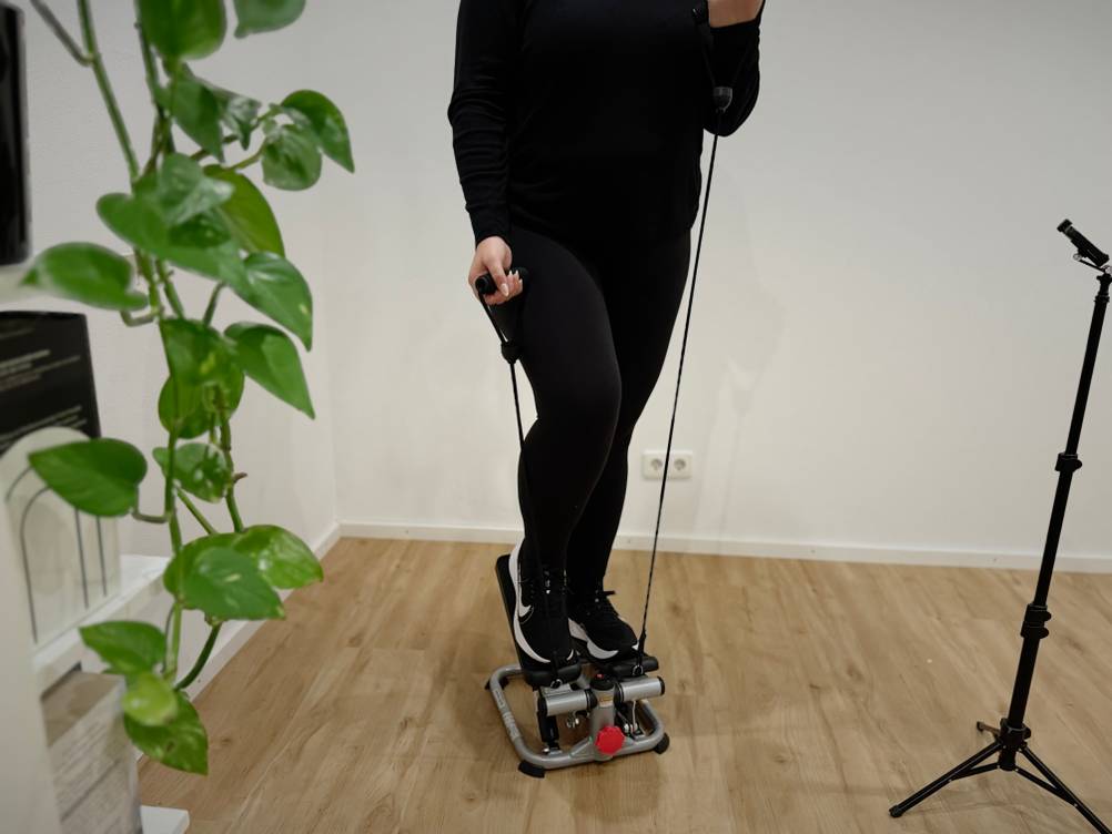 Eine Person in schwarzer Trainingskleidung trainiert auf einem Mini-Stepper und hält die Widerstandsseile fest.