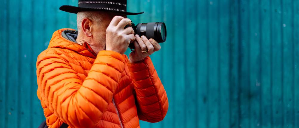 Steppjacke-Herren-Test: Älterer Herr mit schwarzem Hut trägt eine Herren-Steppjacke in Orange beim Fotografieren vor einer blauen Wand.