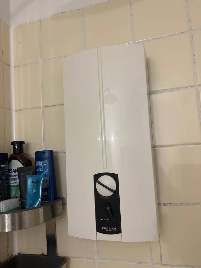 Stiebel Eltron DHB 21 ST Durchlauferhitzer-Test: Das Gerät mit schwarzem Kommandopaneel inklusive weißem Temperaturwahl-Schalter befindet sich an einer hellen Wand neben einem dunklen Regal mit Badeutensilien.