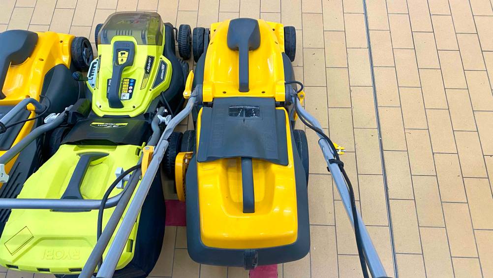 Ansicht von oben auf einen gelb-schwarzen Stiga Elektro-Rasenmäher im Test, der über einen klappbaren Griff verfügt und neben einem grün-schwarzen Akku-Rasenmäher auf beigefarbenen Fliesen steht.