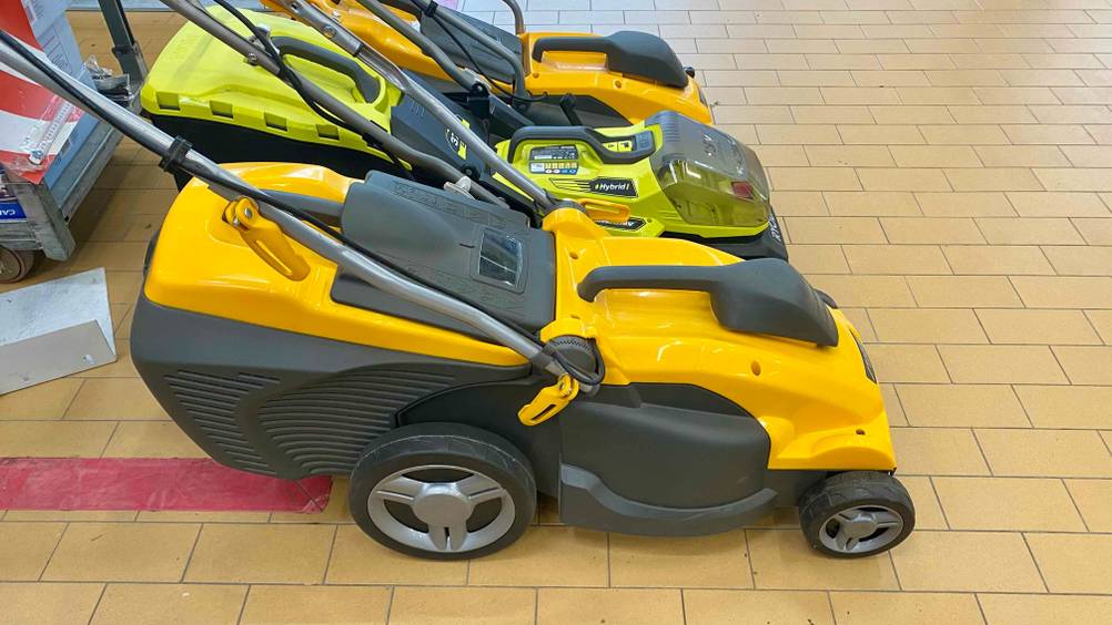 Seitenansicht auf eine Reihe Elektro-Rasenmäher im Test: im Vordergrund ein gelb-schwarzes Stiga Modell inklusive klappbarem Griff auf einem beigefarbenen Fliesenboden.