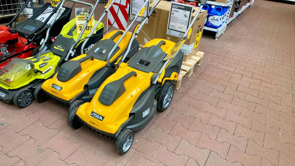 Elektro-Rasenmäher im Test: zwei gelb-schwarze Stiga Modell inklusive klappbarem Griff befinden sich in einer Reihe mit weiteren Rasenmähern auf roten Steinfliesen eines Baumarkts.