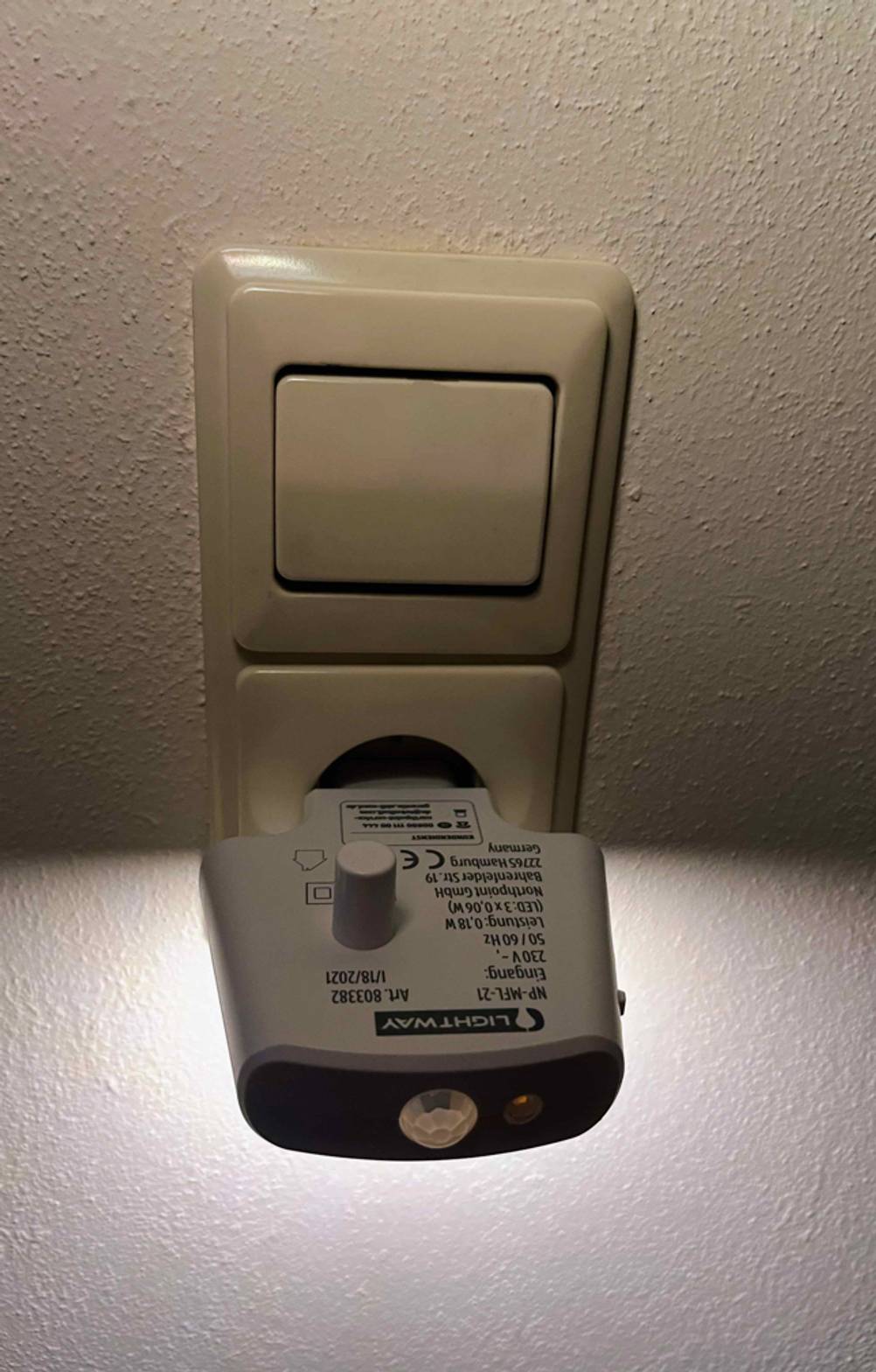 Stilllicht-Test: Ansicht des weißen leuchtenden Steckers, der als Basis und zum Aufladen dient und in einer Steckdose unter einem Lichtschalter steckt.