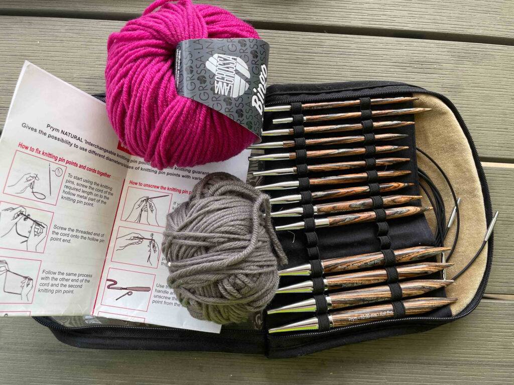 Einige getestete Rund-Stricknadeln mit Holzspitzen befinden sich in ihrer Aufbewahrungstasche, auf der Wolle und eine Gebrauchsanleitung liegen.