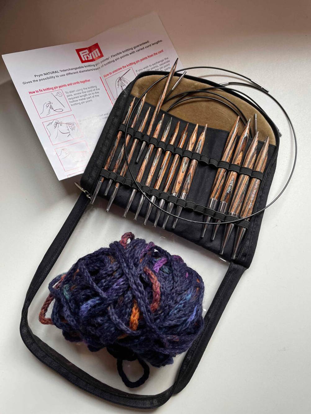 Auf einer hellen Fläche liegt eine Tasche mit unterschiedlichen Stricknadeln im Test, Wolle und einer Bedienungsanleitung.