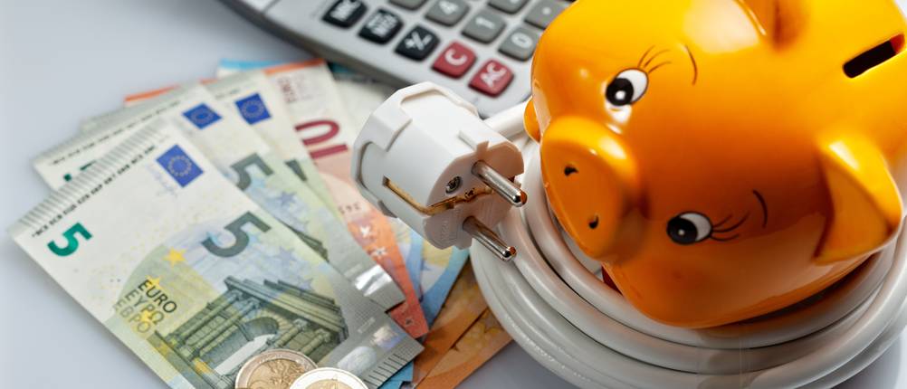 Strom-Test: Auf einem Tisch liegen Geldscheine, Münzen, ein Sparschwein, ein Taschenrechner und ein Stromkabel.