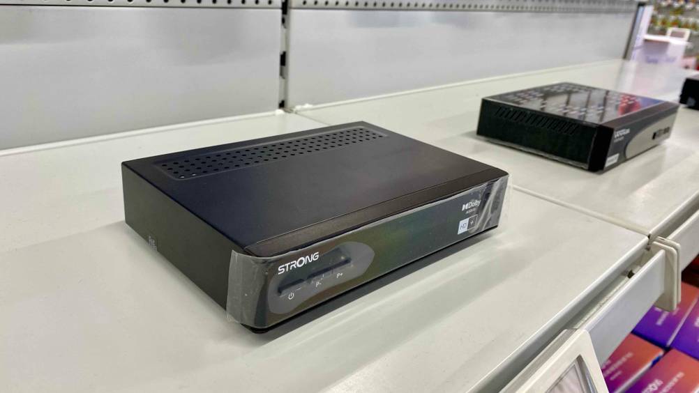 Ein getesteter HD-Sat-Receiver von schräg seitlich auf einem Verkaufregal, im Hintergrund andere Geräte.