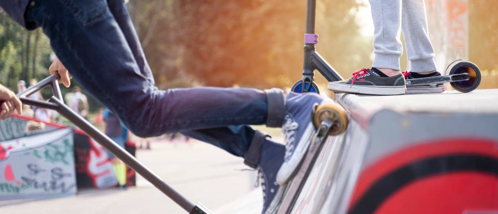 Stunt-Scooter-Test: Person, die mit einem Stunt-Scooter die Halfpipe hinunterfährt, auf dem Scheitelpunkt steht eine weitere Person auf ihrem Scooter