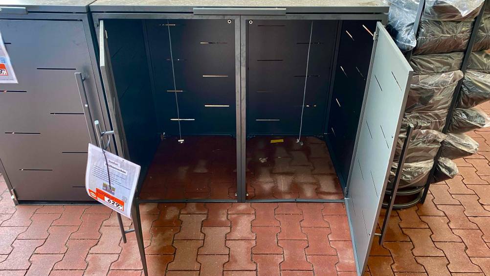 Vorderansicht von schräg oben auf eine Metall-Mülltonnenbox im Test inklusive Preisschild: das graue Modell steht auf einem roten Boden, beide Türen sind geöffnet und zeigen den Platz für zwei Mülltonnen.