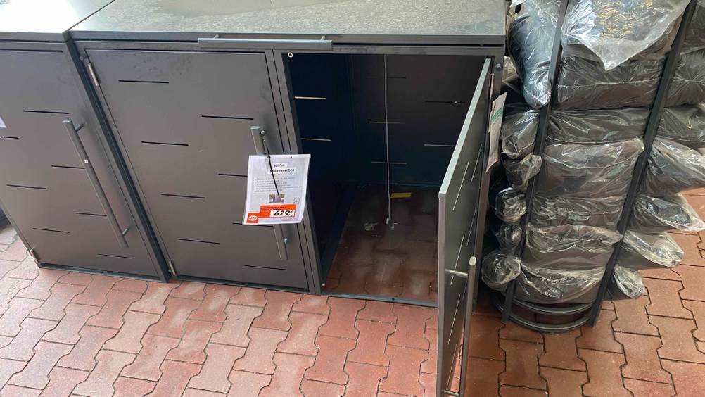 Mülltonnenbox im Test mit Preisschild: ein graues Modell aus Leichtmetall inklusive einer geöffneten Tür, mit Platz für zwei Mülltonnen in einem Ausstellungsbereich auf roten Steinfliesen.