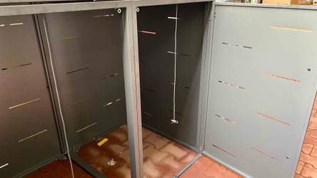 Nahaufnahme einer im Verkaufsbereich auf roten Steinfliesen ausgestellten Mülltonnenbox im Test: aus anthrazitfarbenem Leichtmetall, mit geöffneten Türen und Platz für zwei Mülltonnen.